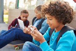 3 Kinder sitzen auf den Boden an einer Mauer und ein Mädchen schaut in einen Laptop wobei ein Junge mit rein schaut und ein Junge auf sein Handy | © monkeybusinessimages - Getty Images/iStockphoto